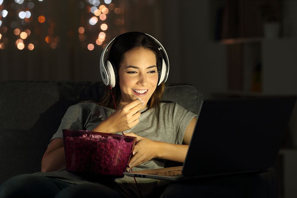 Woman sitting in the dark eating popcorn wearing headphones with laptop in lap watching videos smiling