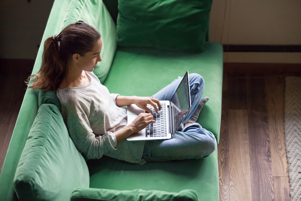 A young woman sits on a green sofa and working on a laptop