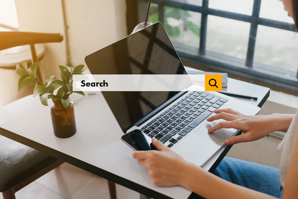 An internet search bar across the screen of a laptop while a woman types at her desk